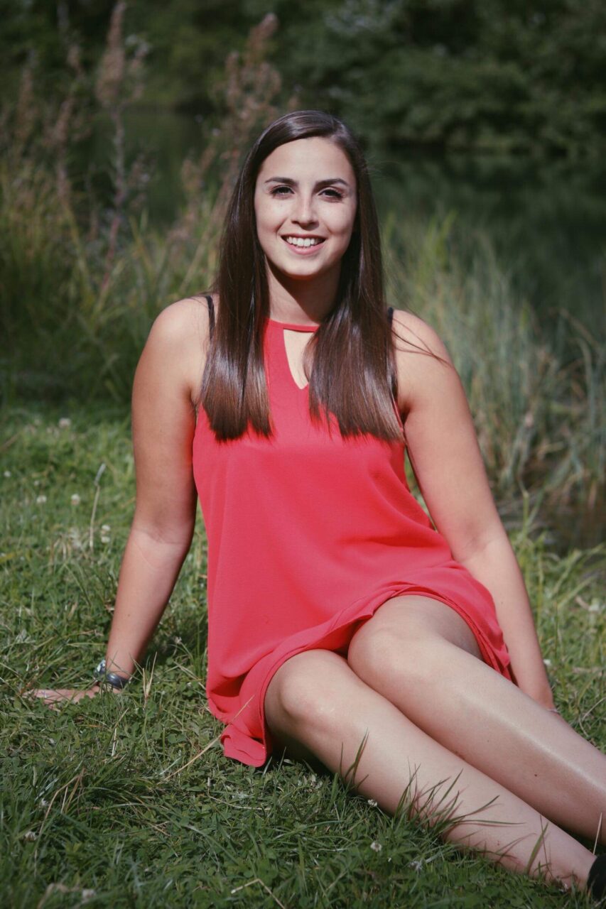 Caroline Mertz en robe corail, assise dans l’herbe (2018)