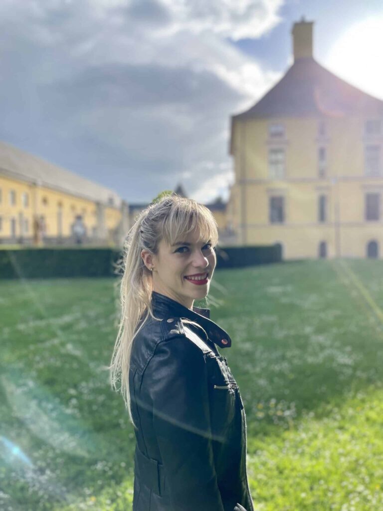 Hélène Henrion en veste de cuir, devant un château aux murs jaunes (2021)
