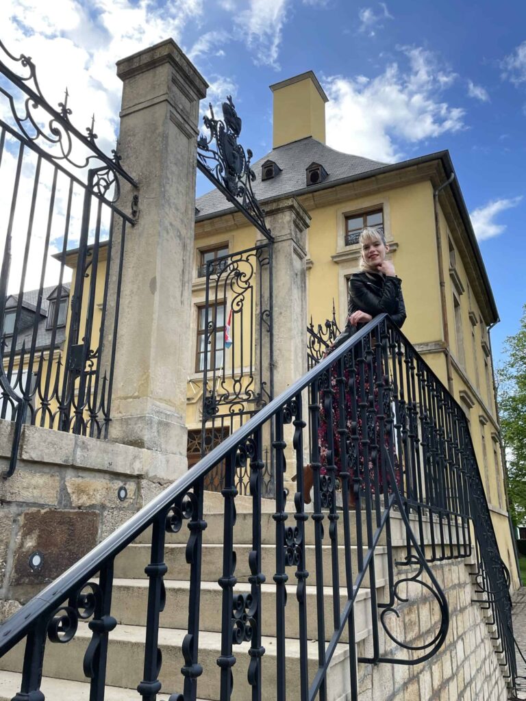 Hélène Henrion sur le perron du château, derrière la grille (2021)