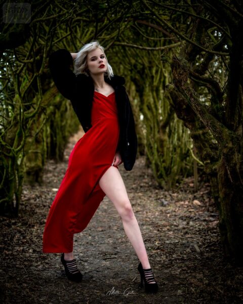 Muriel en robe rouge et manteau noir, sous une voûte d’arbres (photo publiée en février 2022)
