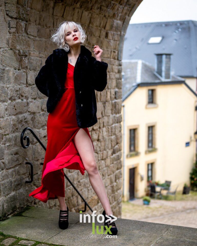 Muriel en robe rouge fendue et manteau de fourrure noir, sous une arche de pierre