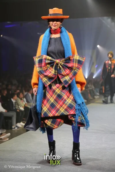 Arlon, Lux Fashion Week 2023. Damlaws, „The new world – Butterfly empire“. Orangefarbener Mantel, große Schleife im Tartanmuster und blauer Schal, orangefarbener Hut