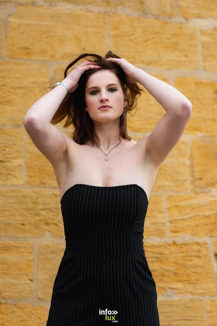 Léanne Stéphano en robe bustier noire, mains dans les cheveux, contre un mur de pierre