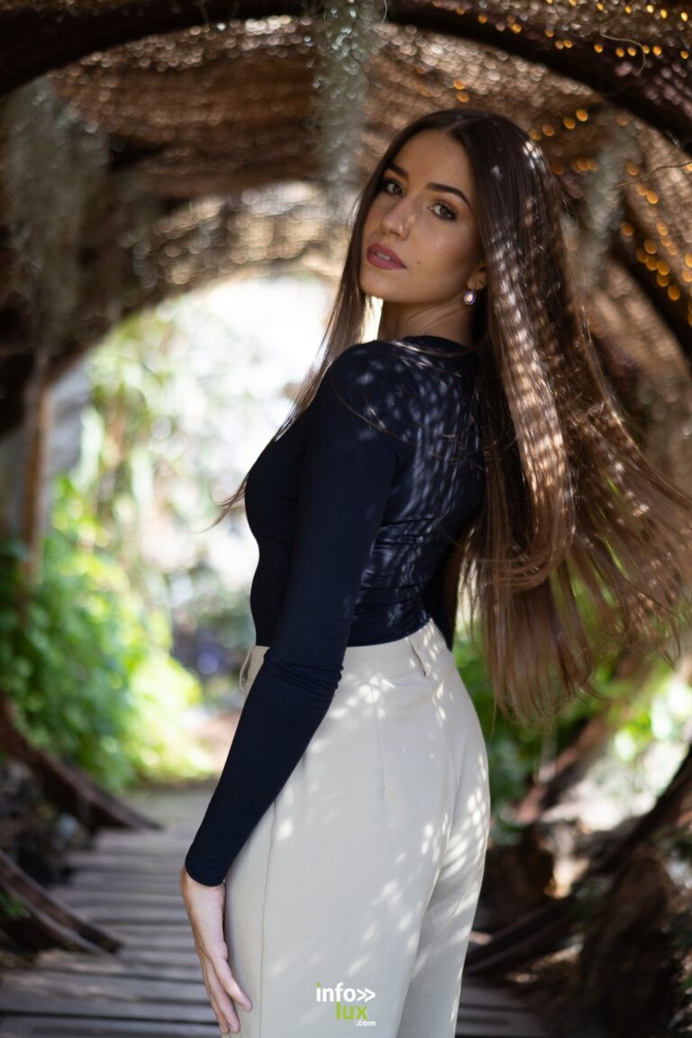 Nina Damblant en haut de dentelle noire et pantalon blanc, sous une arche de verdure