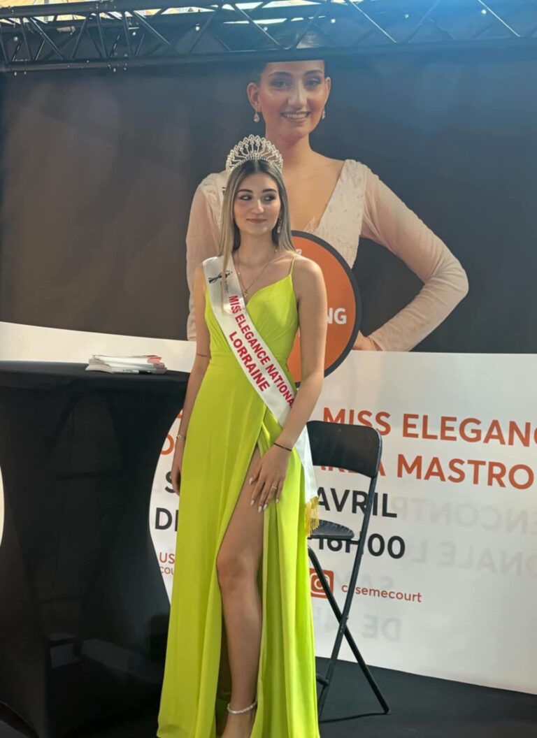 Carla Mastrorillo, couronne et écharpe, devant une affiche annonçant sa venue