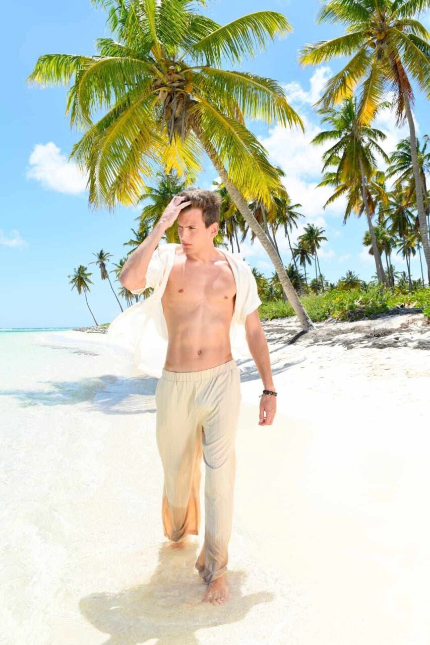 Timothy Legros, chemise ouverte, les pieds dans l’eau d’une plage de sable blanc