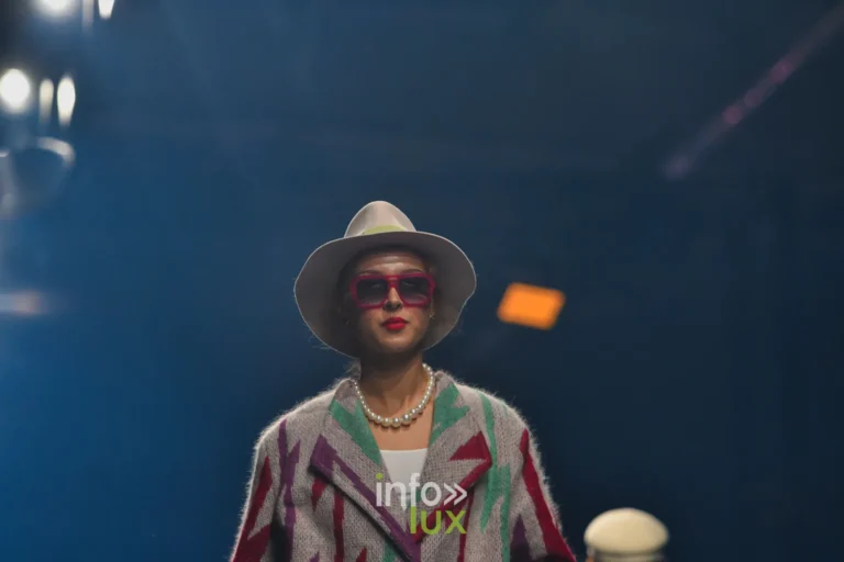 Damlaws, „The Dam Star“. Grauer Strickmantel mit roten, violetten und grünen Blitzmotiven, weißer Hut und rosa Brille
