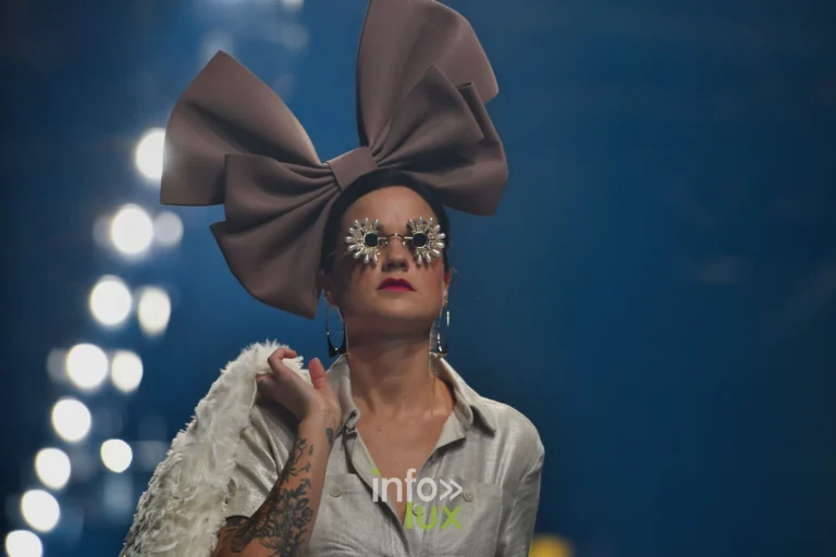 Nœud géant taupe, chemise argentée et étole de plumes blanches — Charlotte Spoidenne, « Diffraction », Lux Fashion Week 2026, Arlon